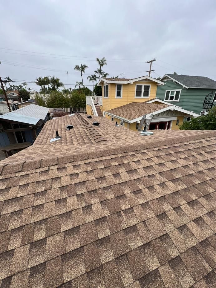 Rooftop view of tan shingle roofs and neighboring houses under a cloudy sky, with palm trees in the background