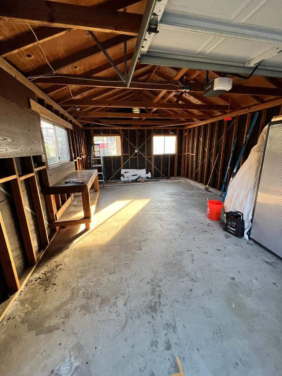 Empty unfinished attic with exposed wooden beams, concrete floor, and sunlight streaming through the windows.