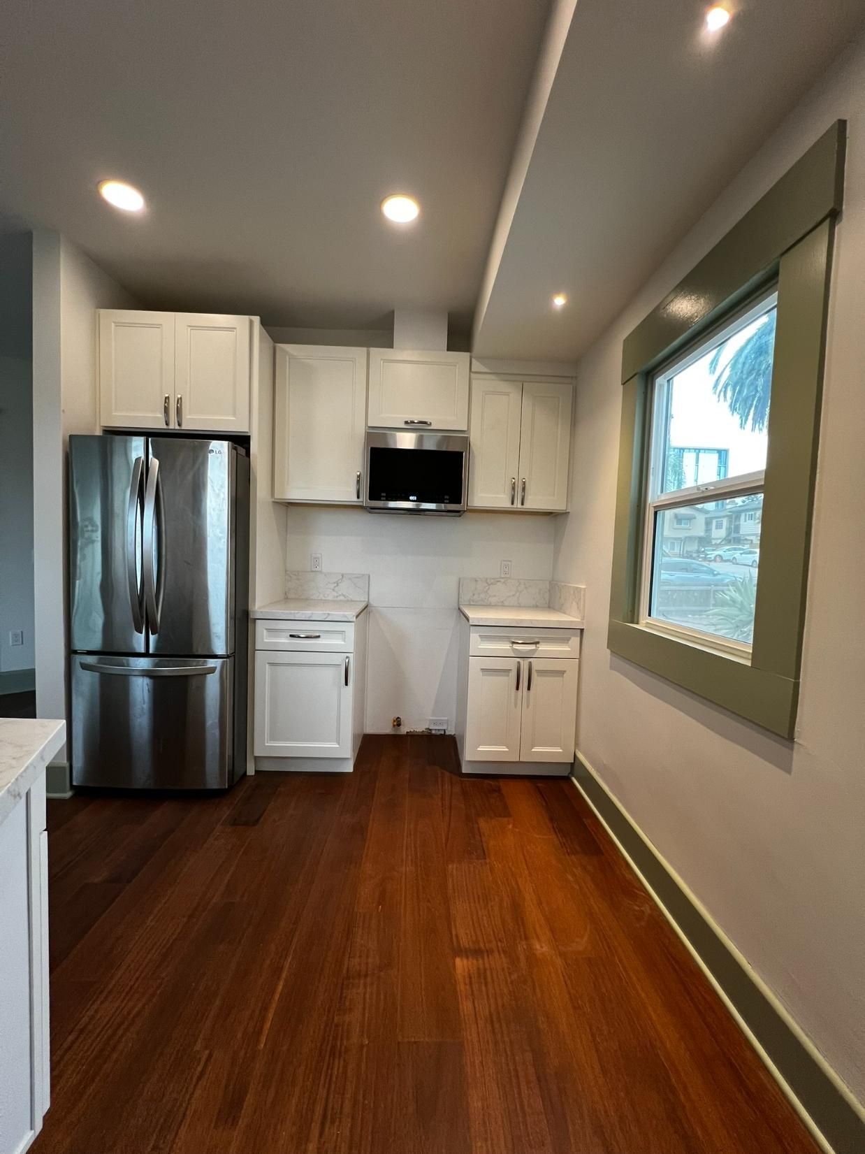 Compact kitchen with white cabinets, stainless fridge, dark wood floors, and a window on the right.