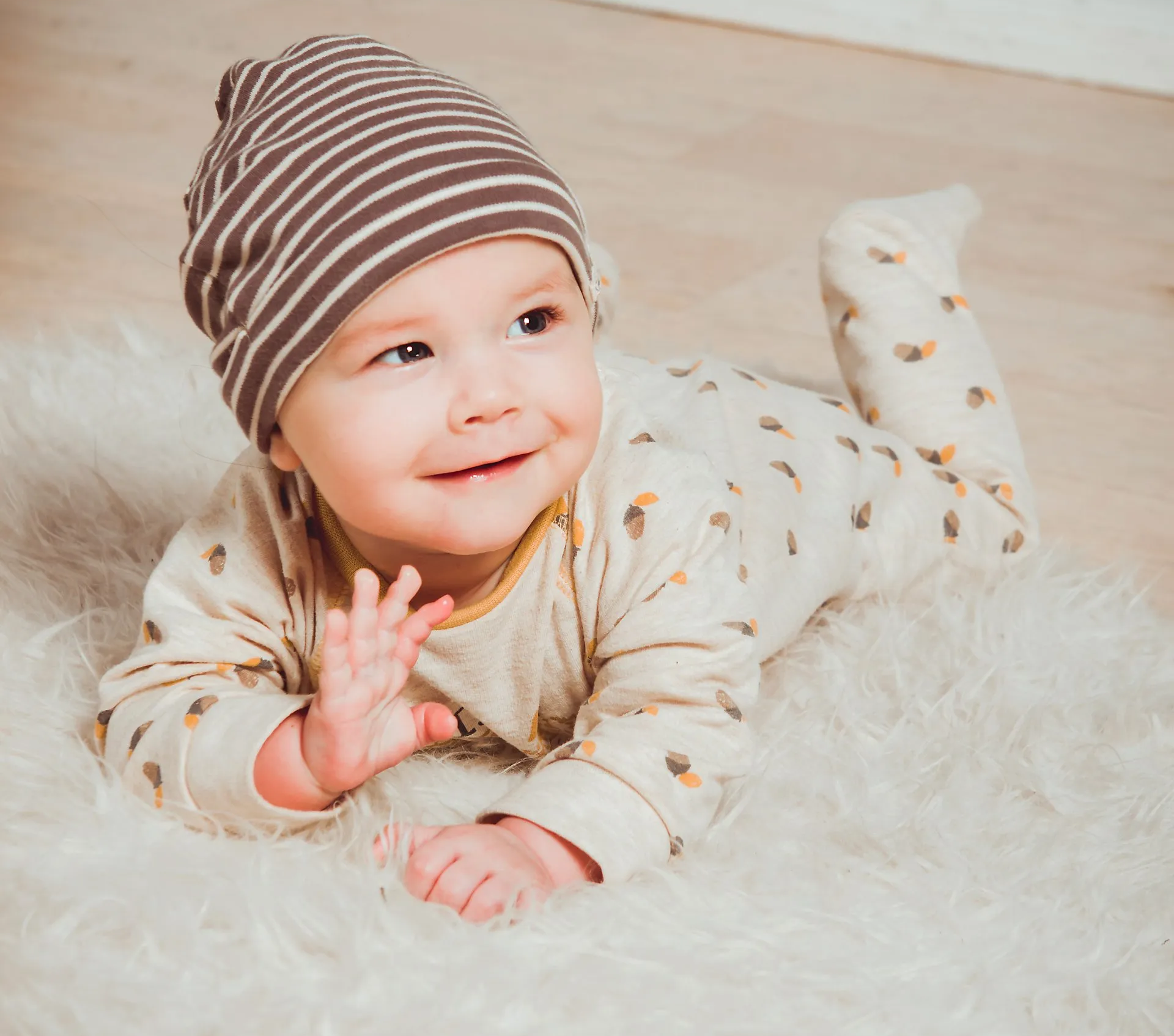 Smiling baby in striped hat and cream outfit лежing on a fluffy white rug.