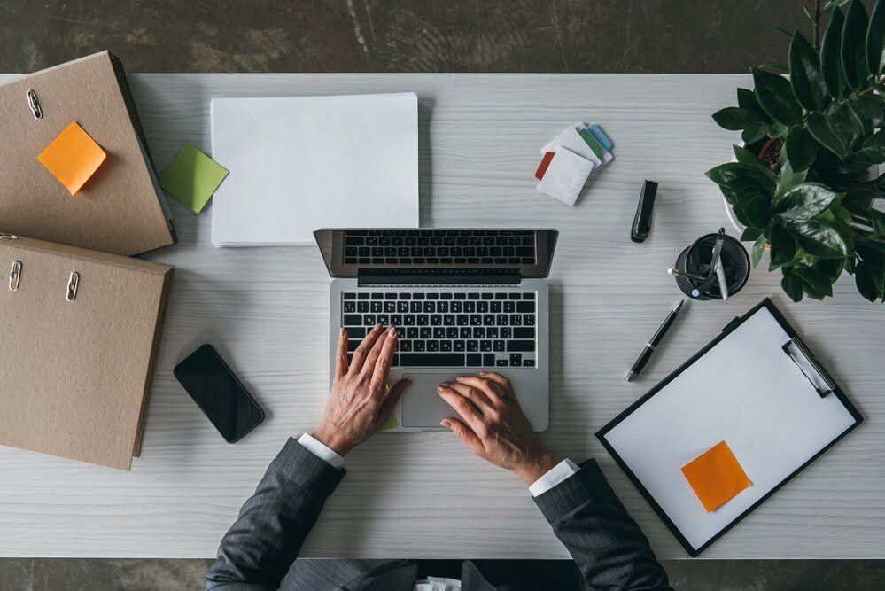 A bird's eye view of a business man working at his desk on his mac, next to a phone, refill pad, binders, and pens.