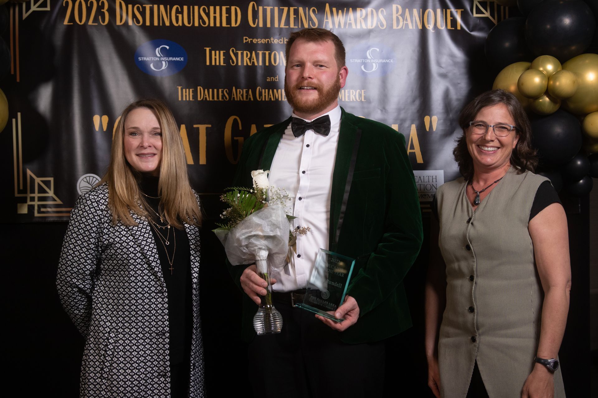 A man in a tuxedo is holding an award while standing next to two women.