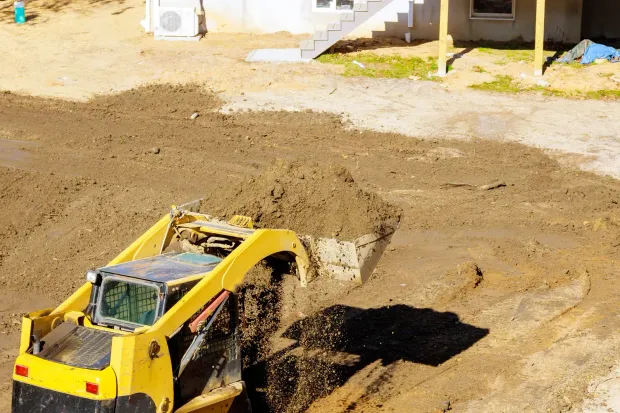 Yellow excavator working on a muddy construction site beside a brick wall and shallow water.
