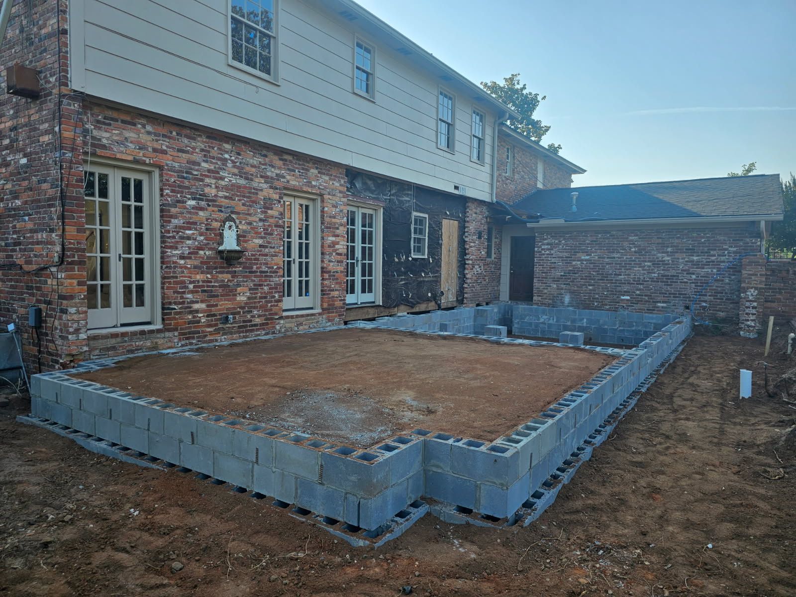 Backyard patio under construction beside brick and siding house, with concrete block border and dirt base