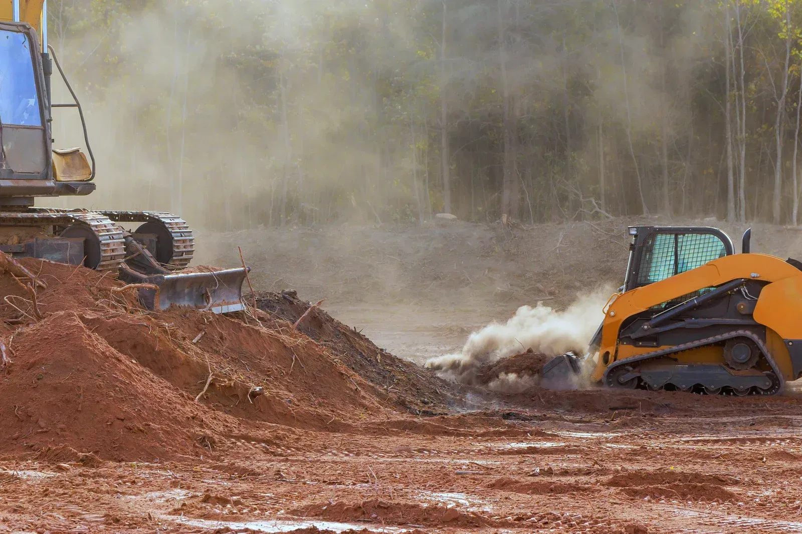 Excavators moving red dirt at a muddy construction site, with dust cloud and trees in the background