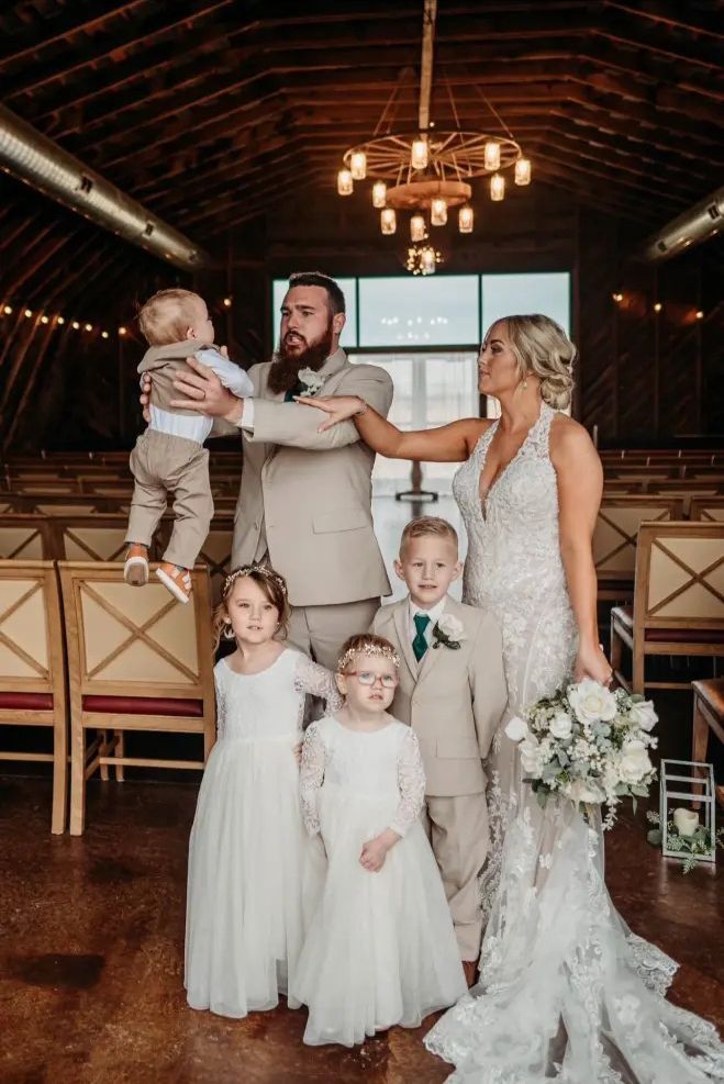 Wedding party indoors. Bride in white dress, groom in tan suit, holding a child. Children in formal wear.