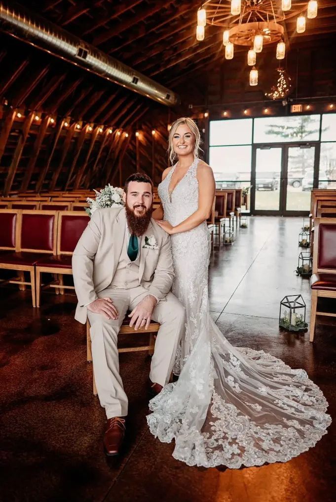 Bride and groom pose inside a barn. The bride wears a white gown with lace train. The groom in a tan suit.
