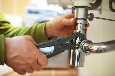 Hands using a wrench to tighten a chrome pipe under a sink.