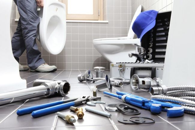 Plumbing tools scattered on a bathroom floor beside a toilet and sink, with a person standing nearby.