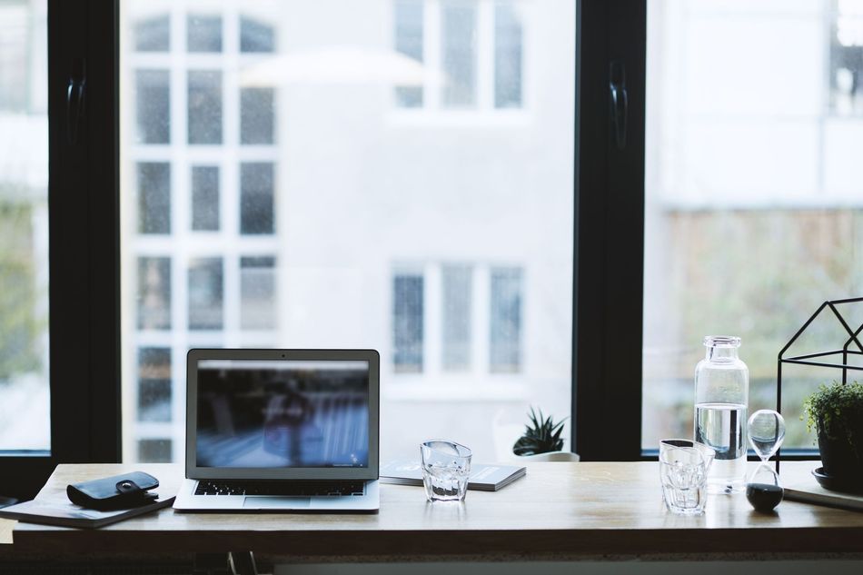 Laptop on a desk by a bright window with a water bottle, glasses, and small plants