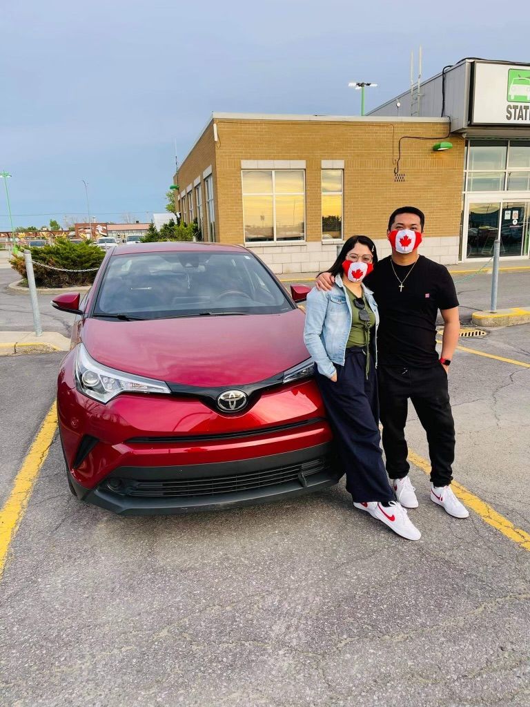 A man and a woman wearing masks are standing in front of a red car.
