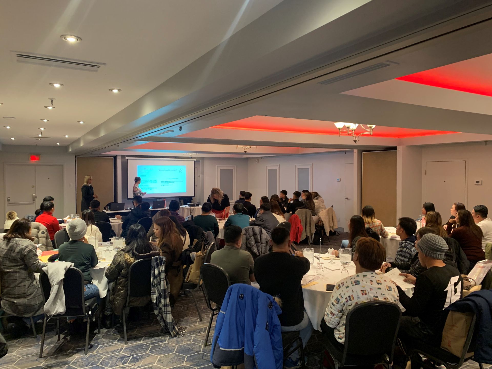 A group of people are sitting at tables in a large room watching a presentation.