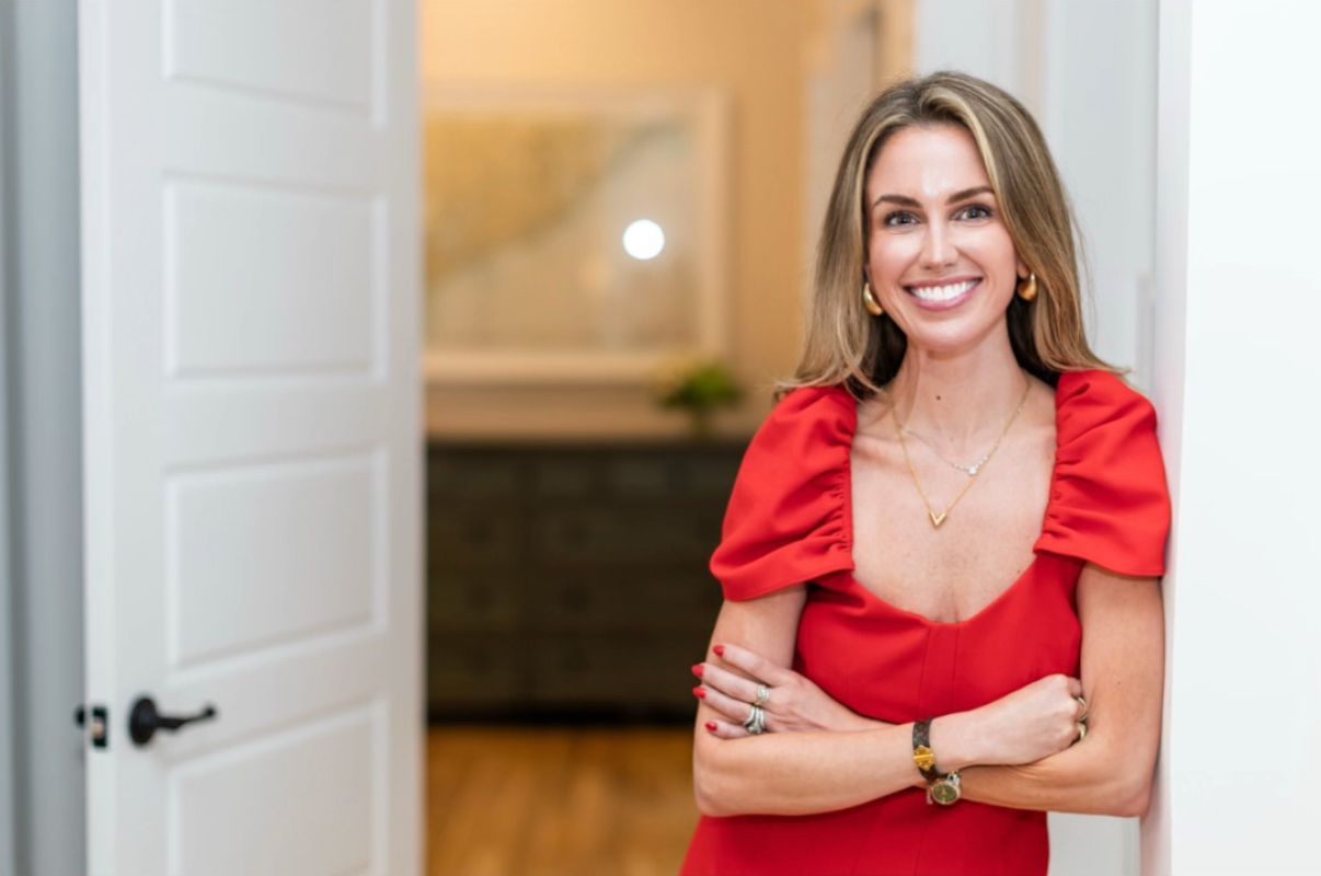 Ashley Cashon in a red dress, leaning against a doorway with arms crossed, smiling inside a home, featured on About Me page