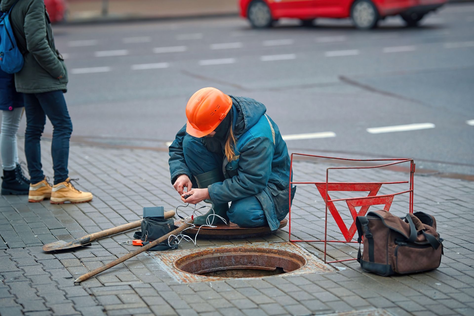 Worker in orange helmet repairing a manhole on a city sidewalk, with tools and a safety sign nearby