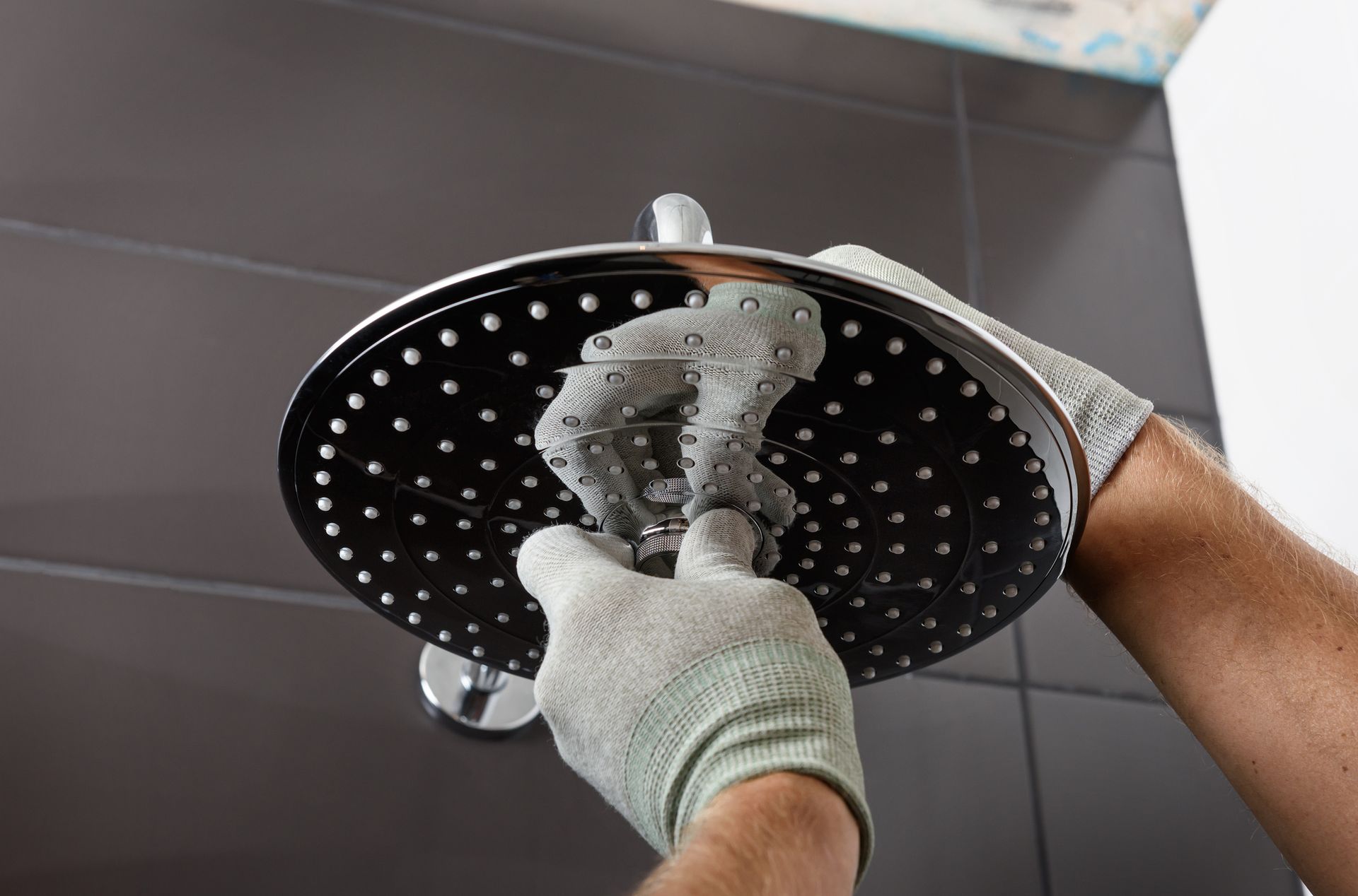 A person wearing work gloves is cleaning a black showerhead with a sponge.