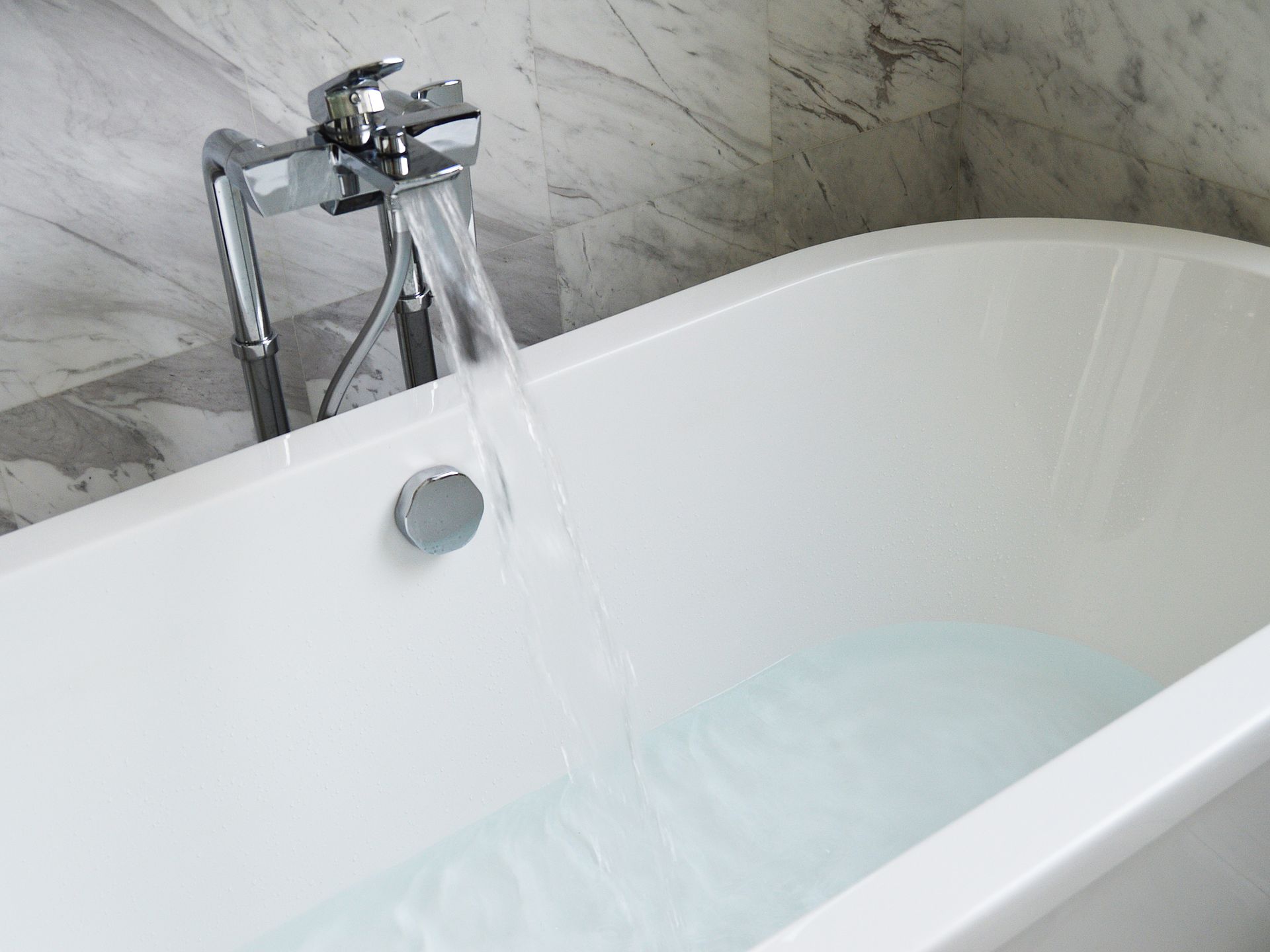 White bathtub filling with water from a chrome faucet, against a marble wall.