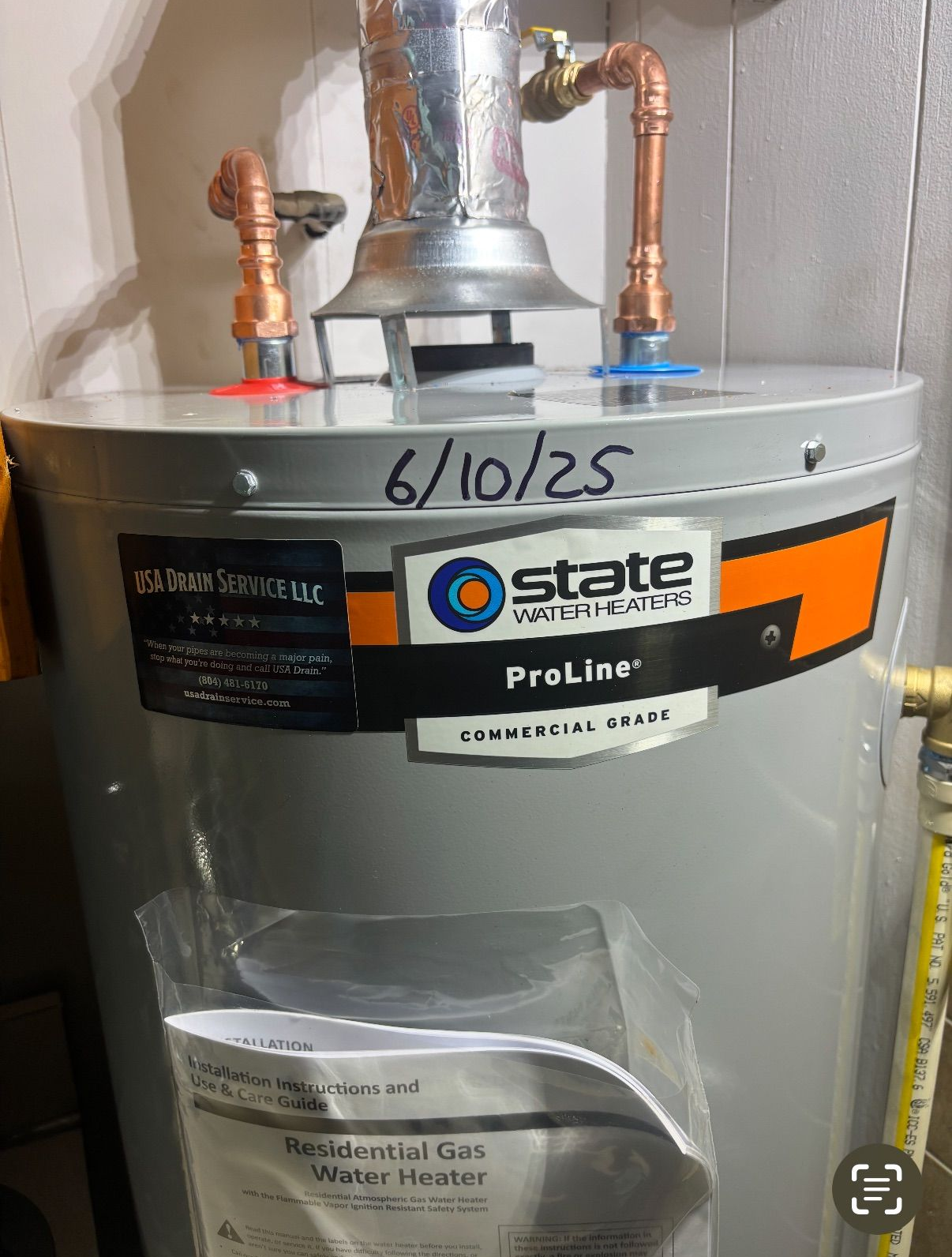 White and black water heater system in a room, pipes and boxes against a wall.
