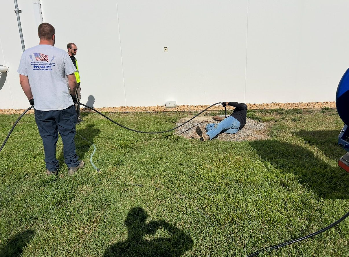 A worker in blue overalls and gloves uses a sewer snake on an open manhole in a brick paved area.