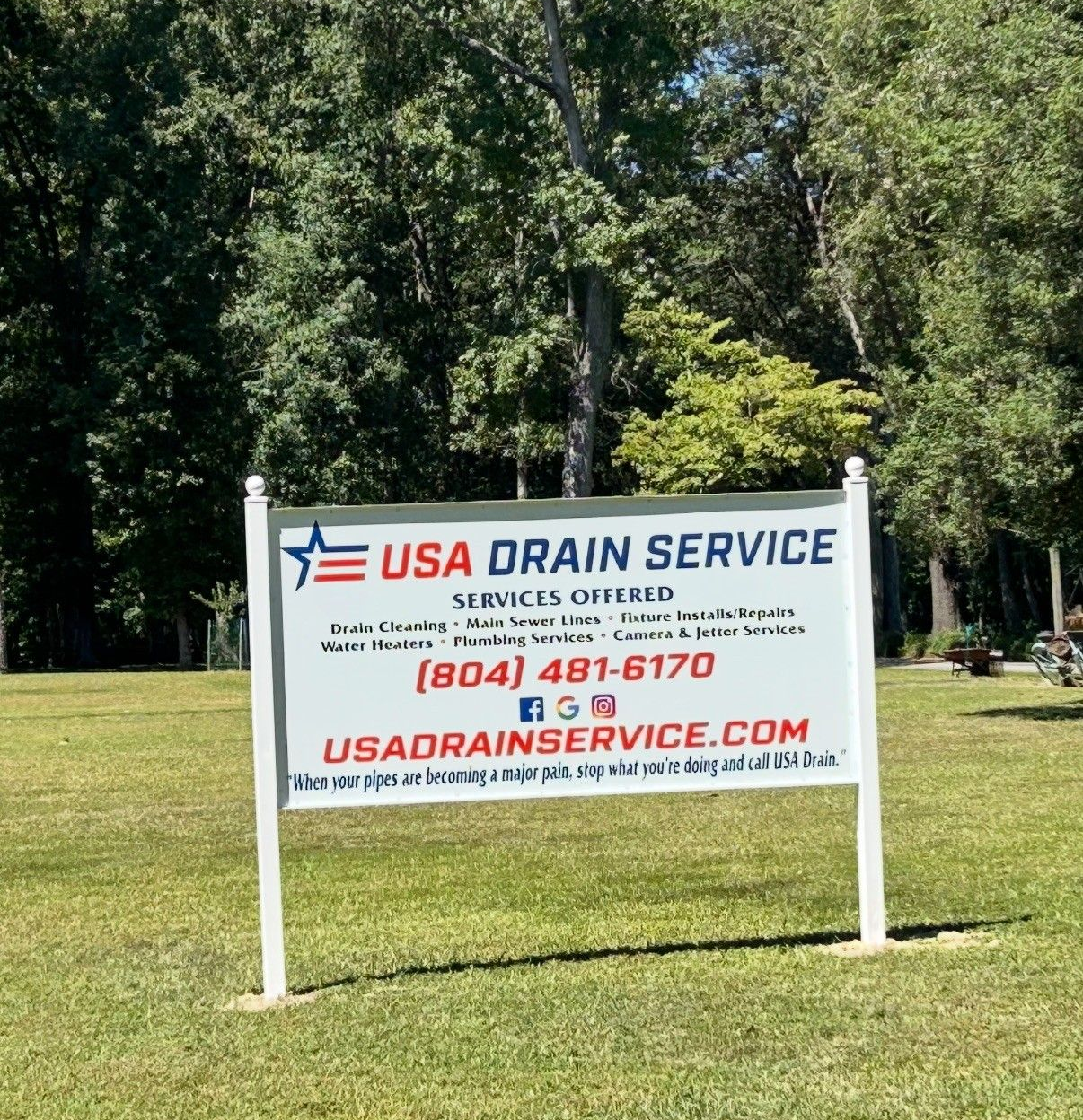 USA Drain Service sign outdoors in a grassy area with trees in the background; white sign with blue and red text.