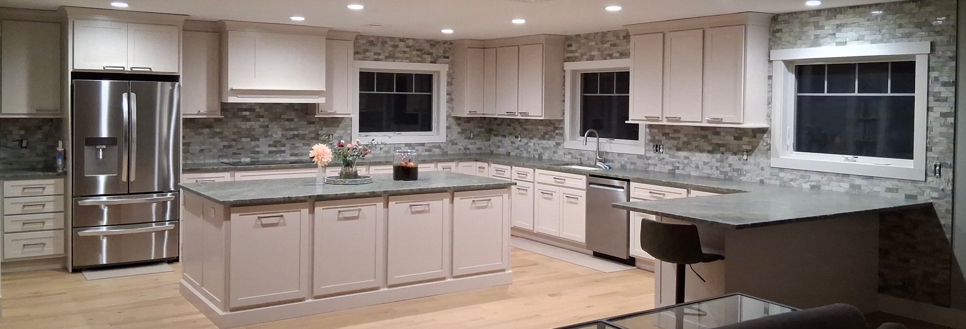 Modern white kitchen with island, stainless steel fridge, marble countertops, and large window over the sink.