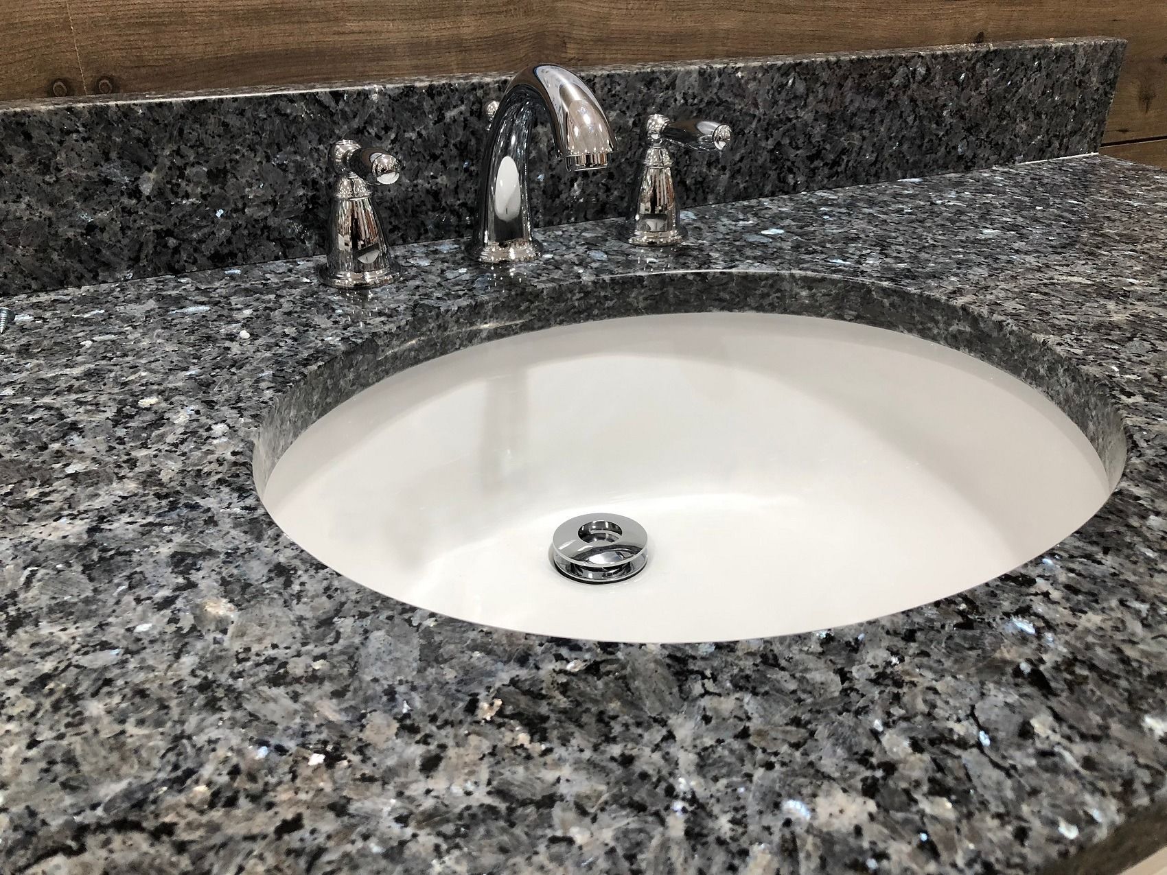 White oval bathroom sink set in speckled granite countertop with chrome faucet and drain.