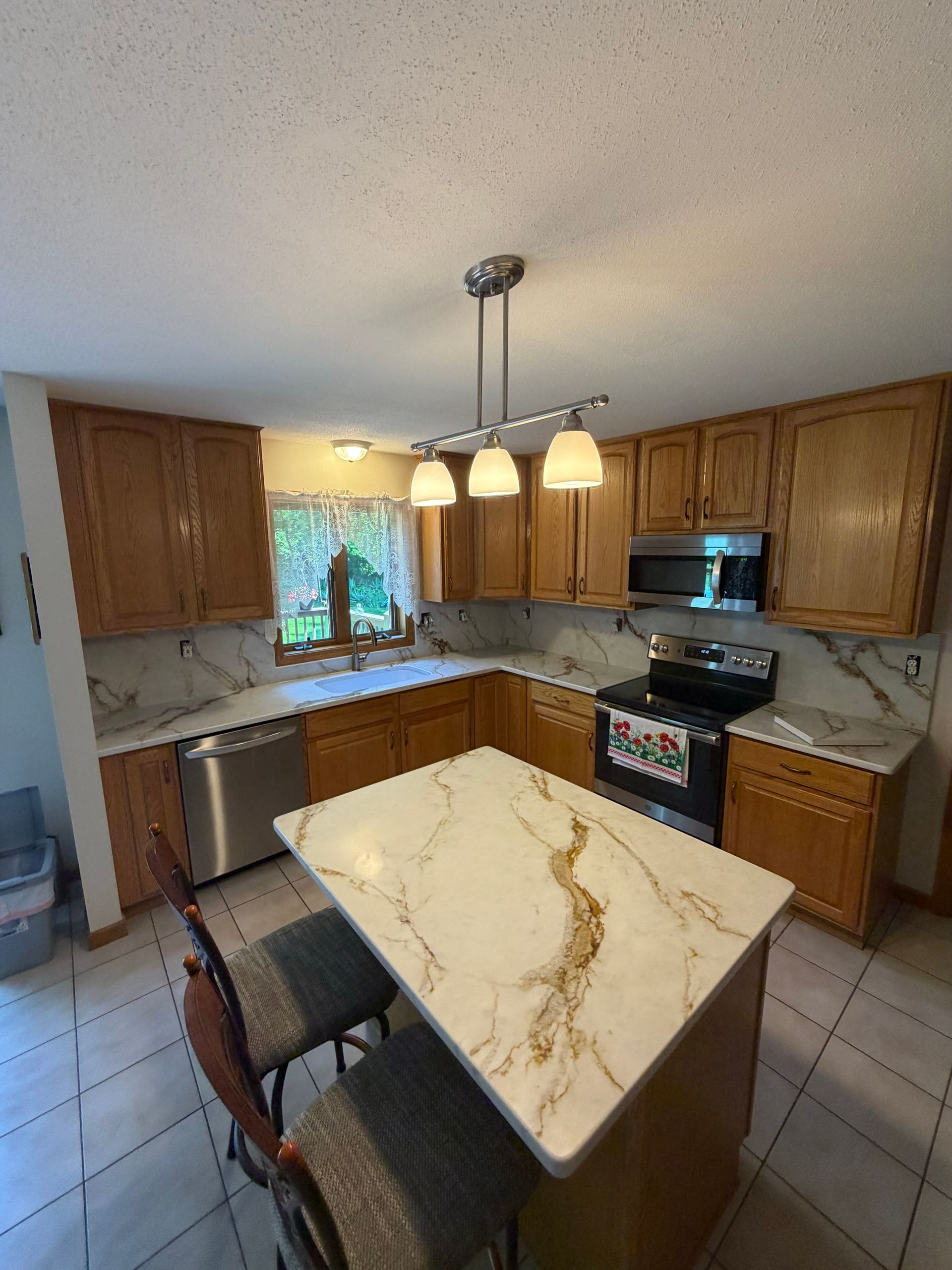 Bright kitchen with marble island, wooden cabinets, stainless appliances, and a hanging pendant light.