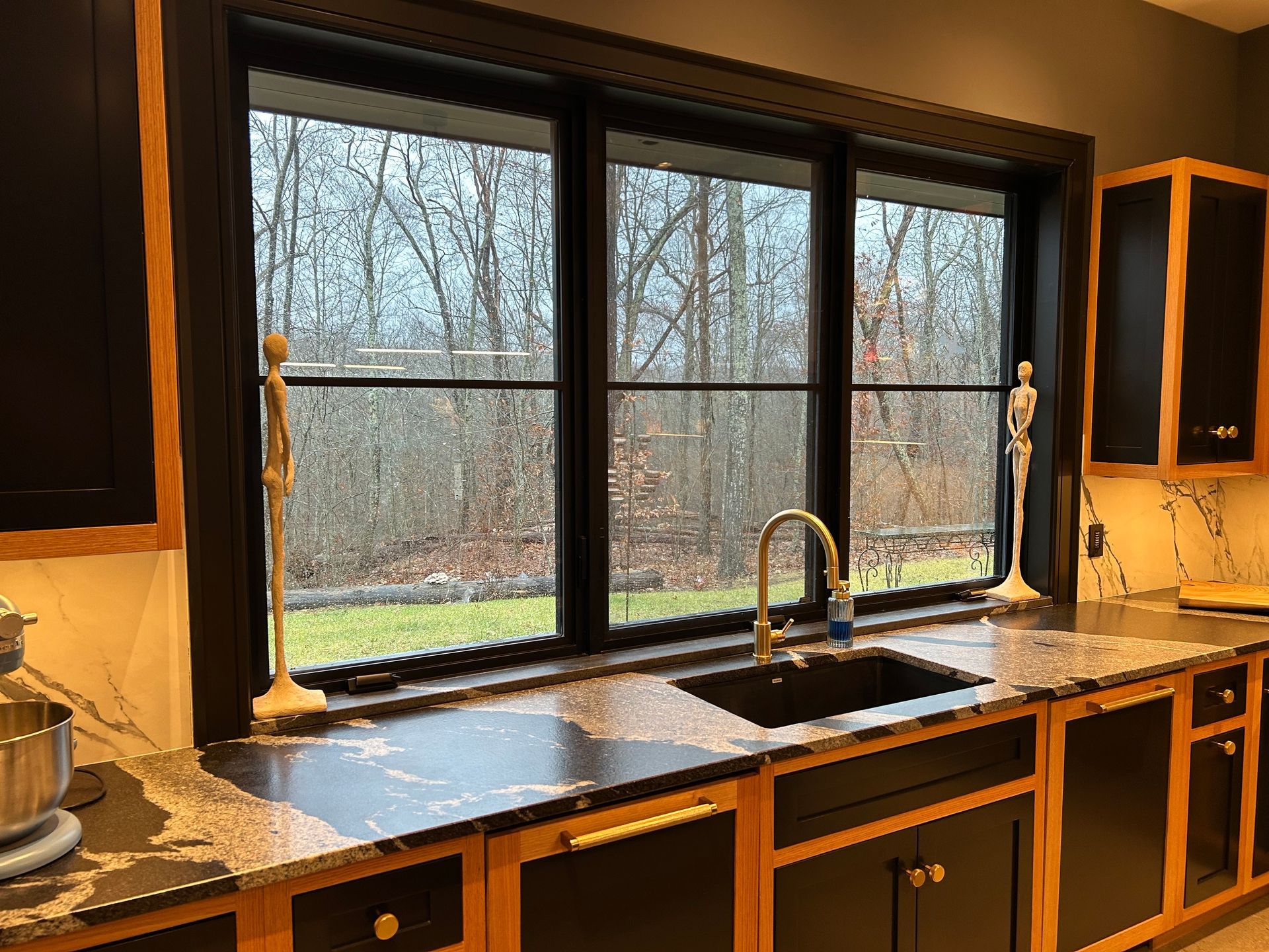 Kitchen sink beneath a large window, with dark countertops, wooden cabinets, and a wooded view outside.