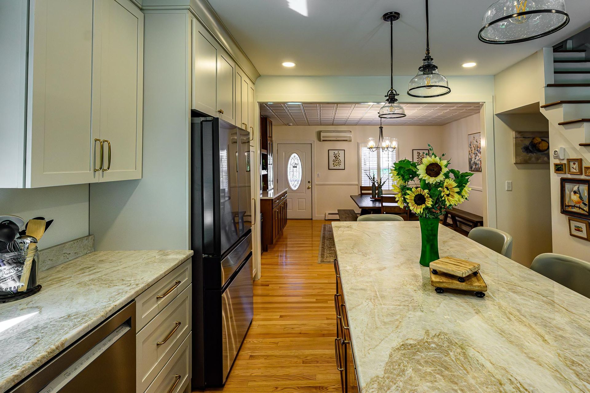 Bright kitchen with white cabinets, granite island, wooden floor, stainless fridge, and sunflowers on the counter