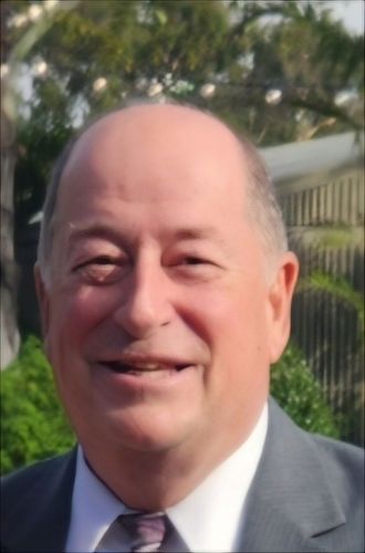 Man in a navy suit and white shirt smiles, indoors with a blurred background.