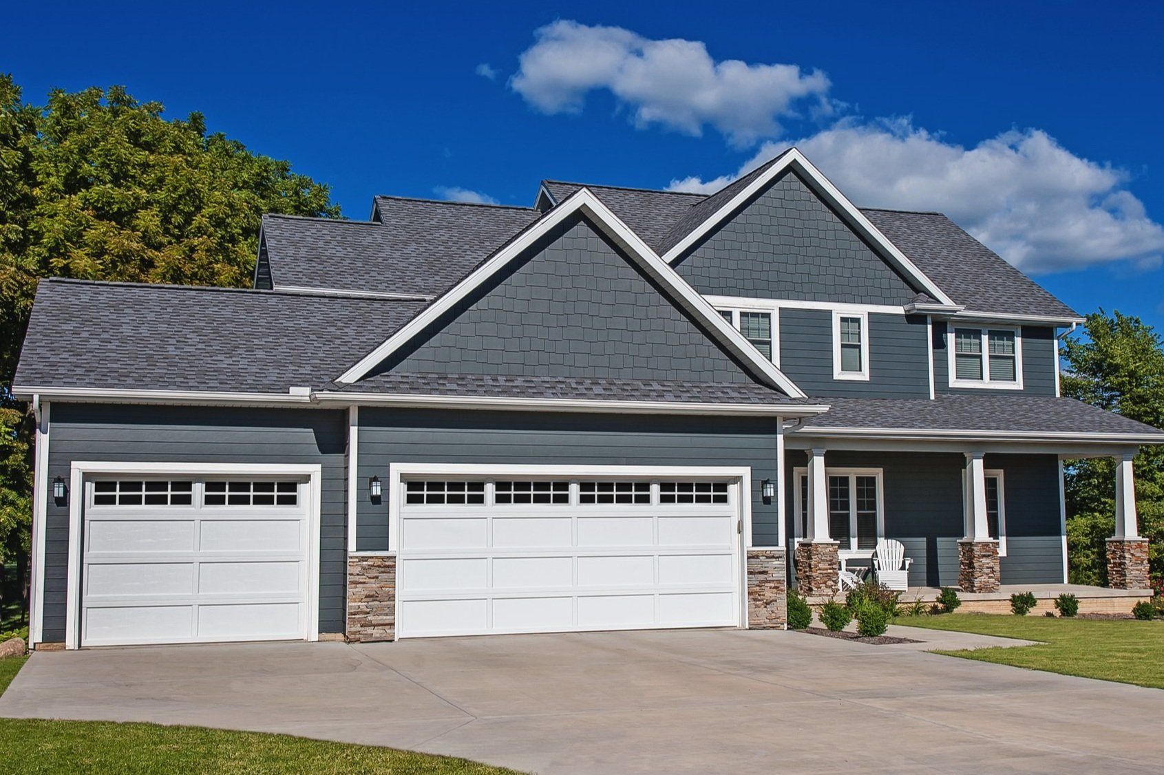 Two-story blue house with white garage doors and a porch, set against a blue sky with clouds.