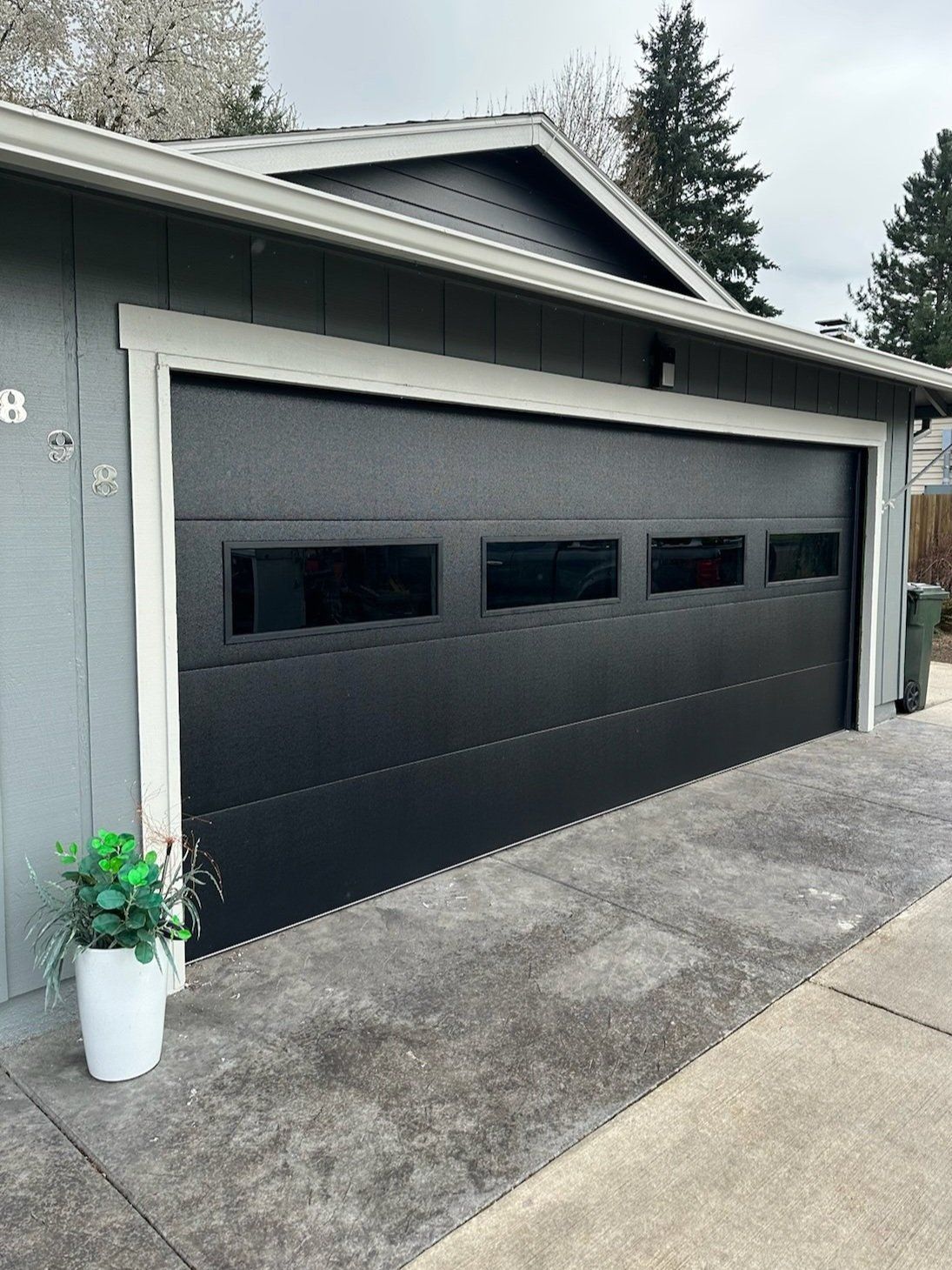 Black garage door with rectangular windows, gray siding, concrete driveway, white trim.