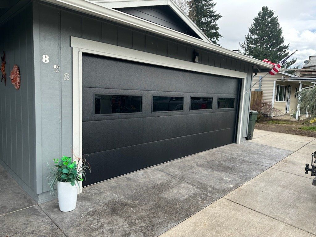Black garage door with rectangular windows, gray siding, concrete driveway, and a potted plant.