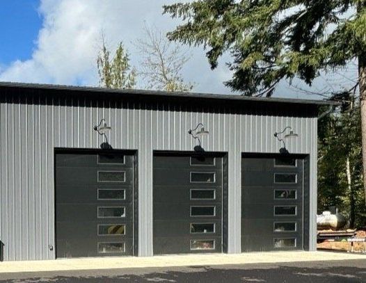 Three-bay garage with black doors and glass panels. Exterior metal siding, overhead lights, and a tree background.