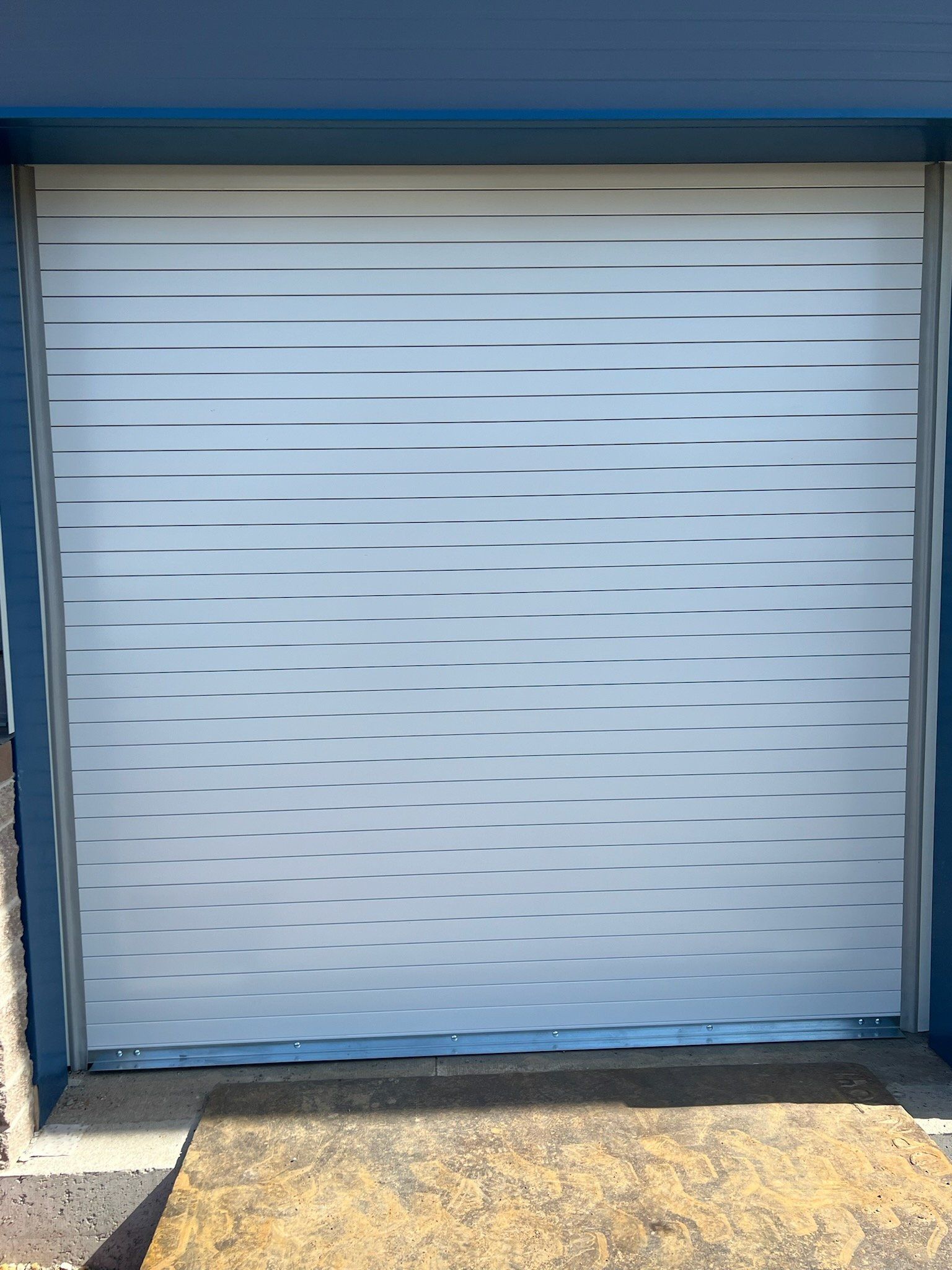 White roll-up door with wavy pattern on a blue building. A concrete ramp is in front.