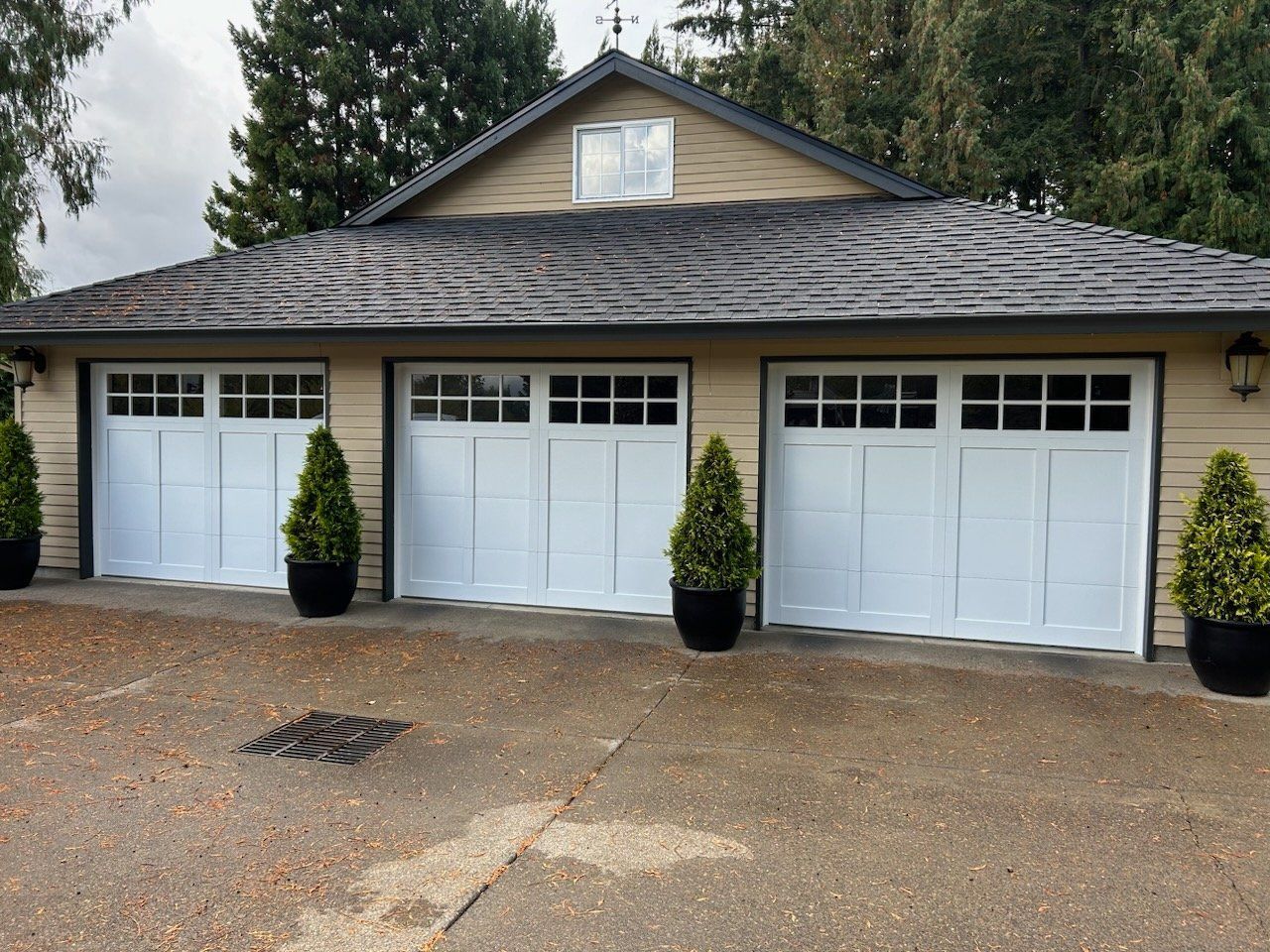 Three white garage doors with black trim under a textured roof. Black potted plants flank the doors.
