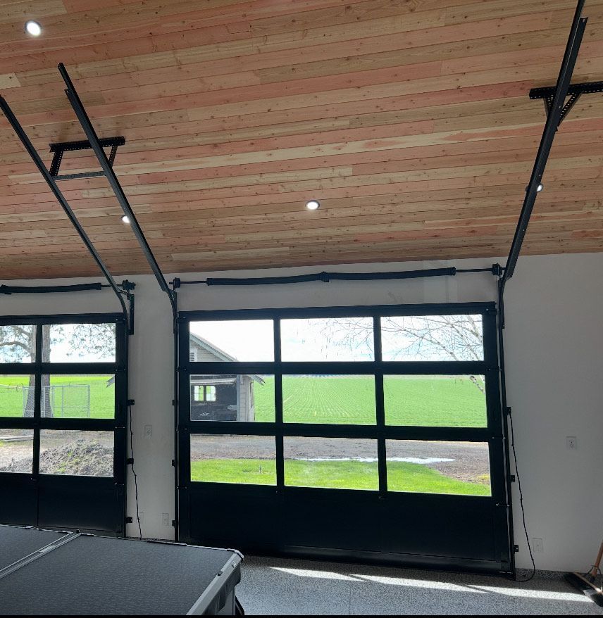 Two black garage doors with glass panels, open to a green field. Light wooden ceiling.
