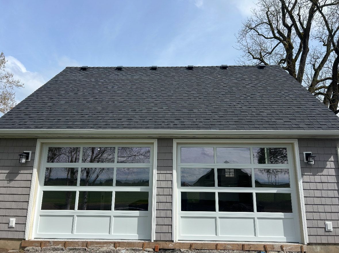 Two glass-paned garage doors with white frames on a gray-shingled building, beneath a dark shingle roof, with a blue sky.