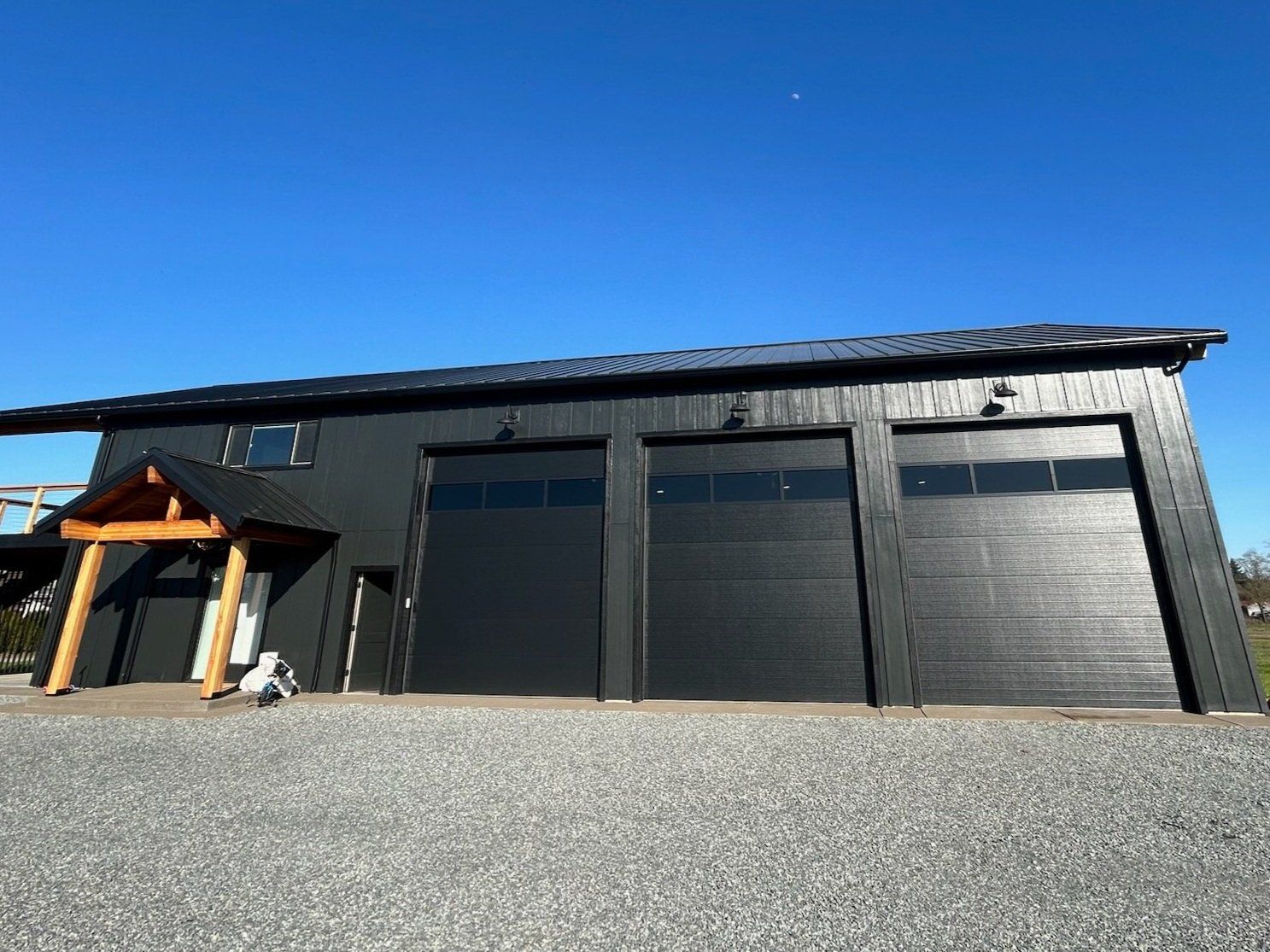 Black garage with three bays and a small wooden entry under a blue sky.