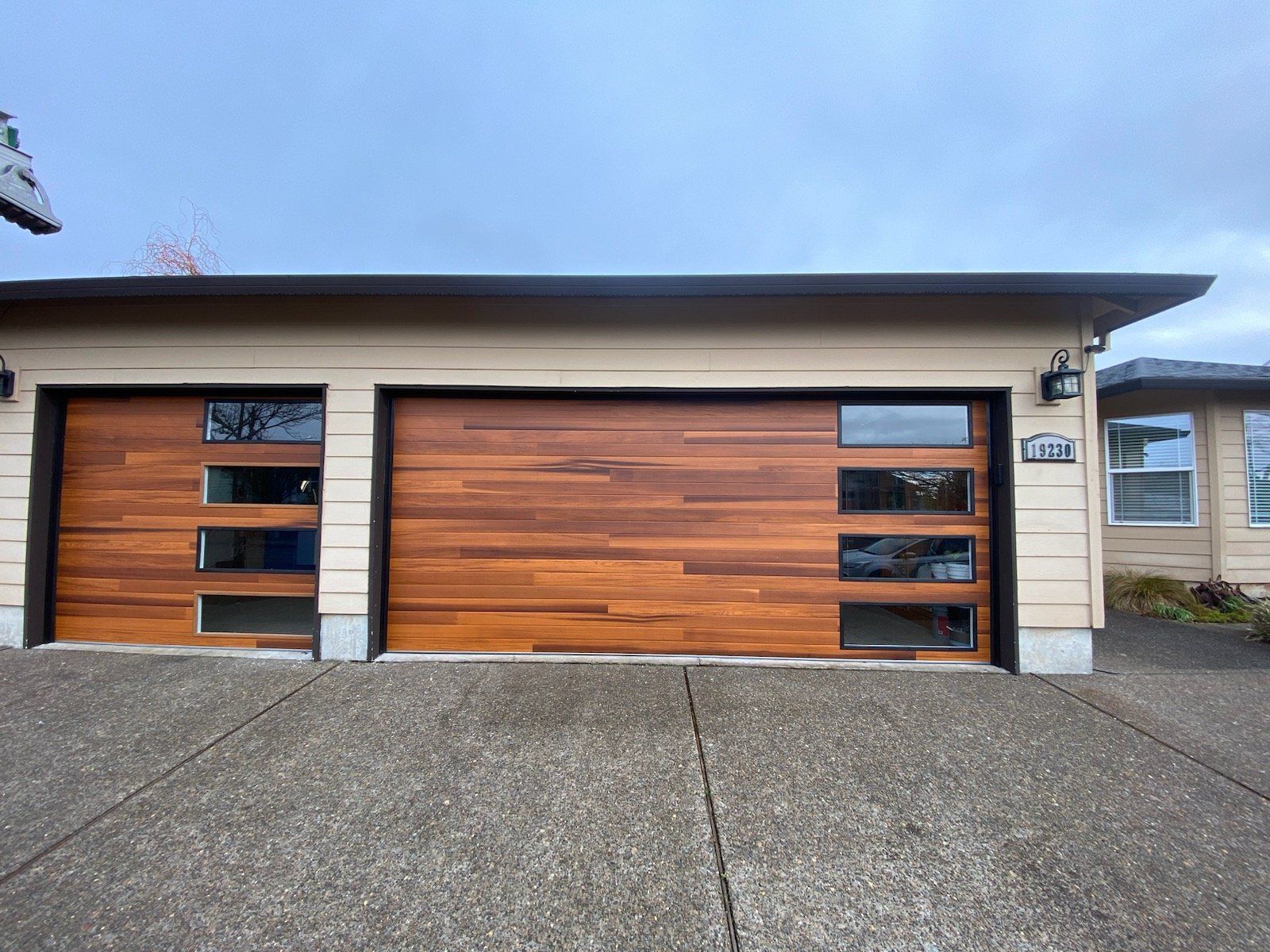 Two-car garage with wood grain doors and rectangular windows. Tan exterior, gray concrete driveway, overcast sky.