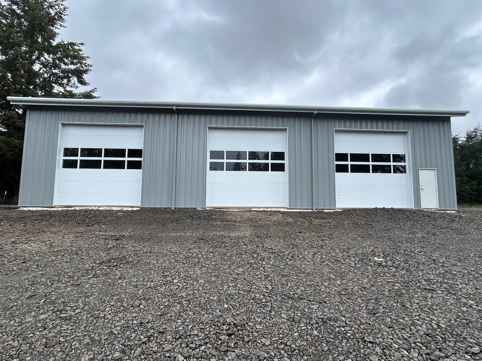 Gray metal building with three white garage doors on a gravel lot under a cloudy sky.