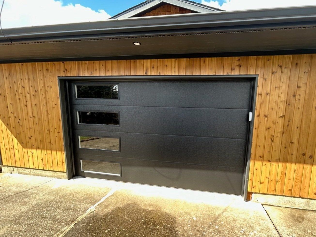 Black garage door with glass panels set in a wooden building, against a blue sky.