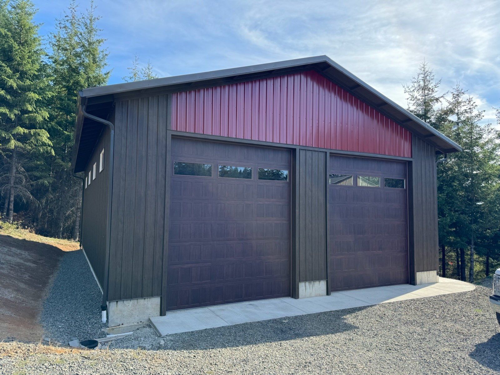 Two-car garage with dark brown siding and maroon roof, built on a concrete foundation in a wooded area.