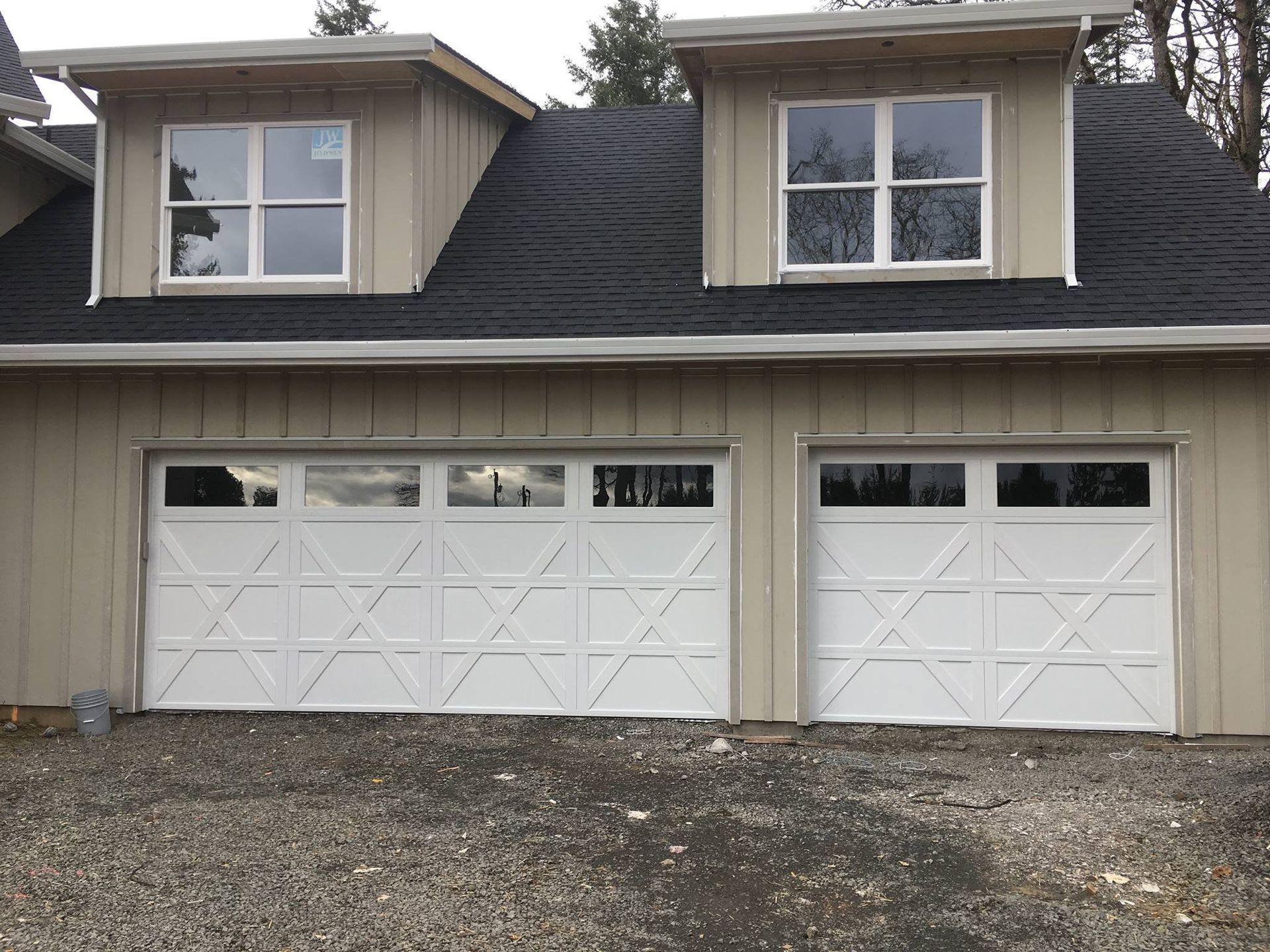 Two-car garage with white doors, tan siding, black roof, and two small dormers with windows.