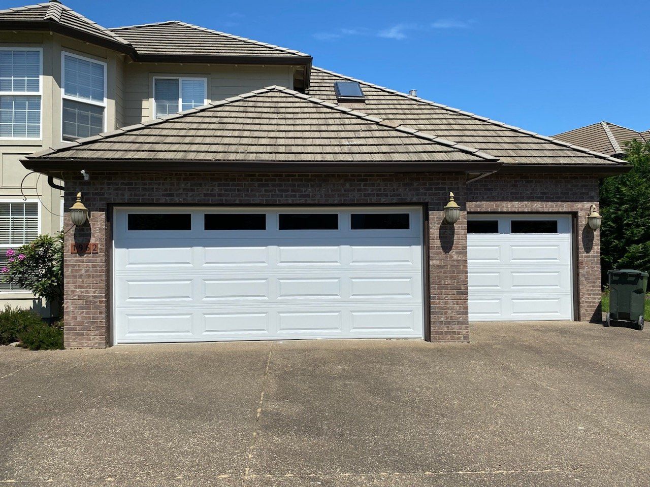 Two-car garage with white doors, brick facade, and brown shingled roof under a clear, blue sky.