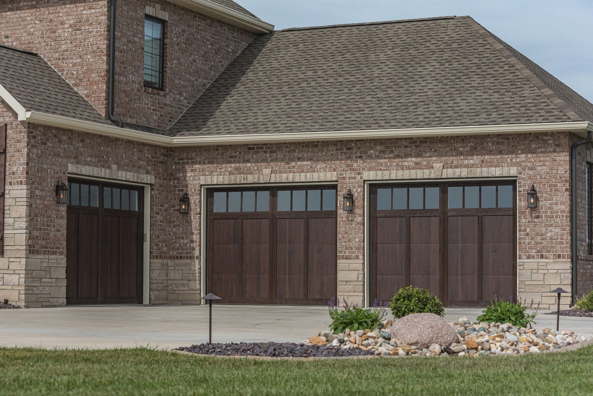 Three-car garage with brown doors, brick exterior, and a stone garden on the lawn.