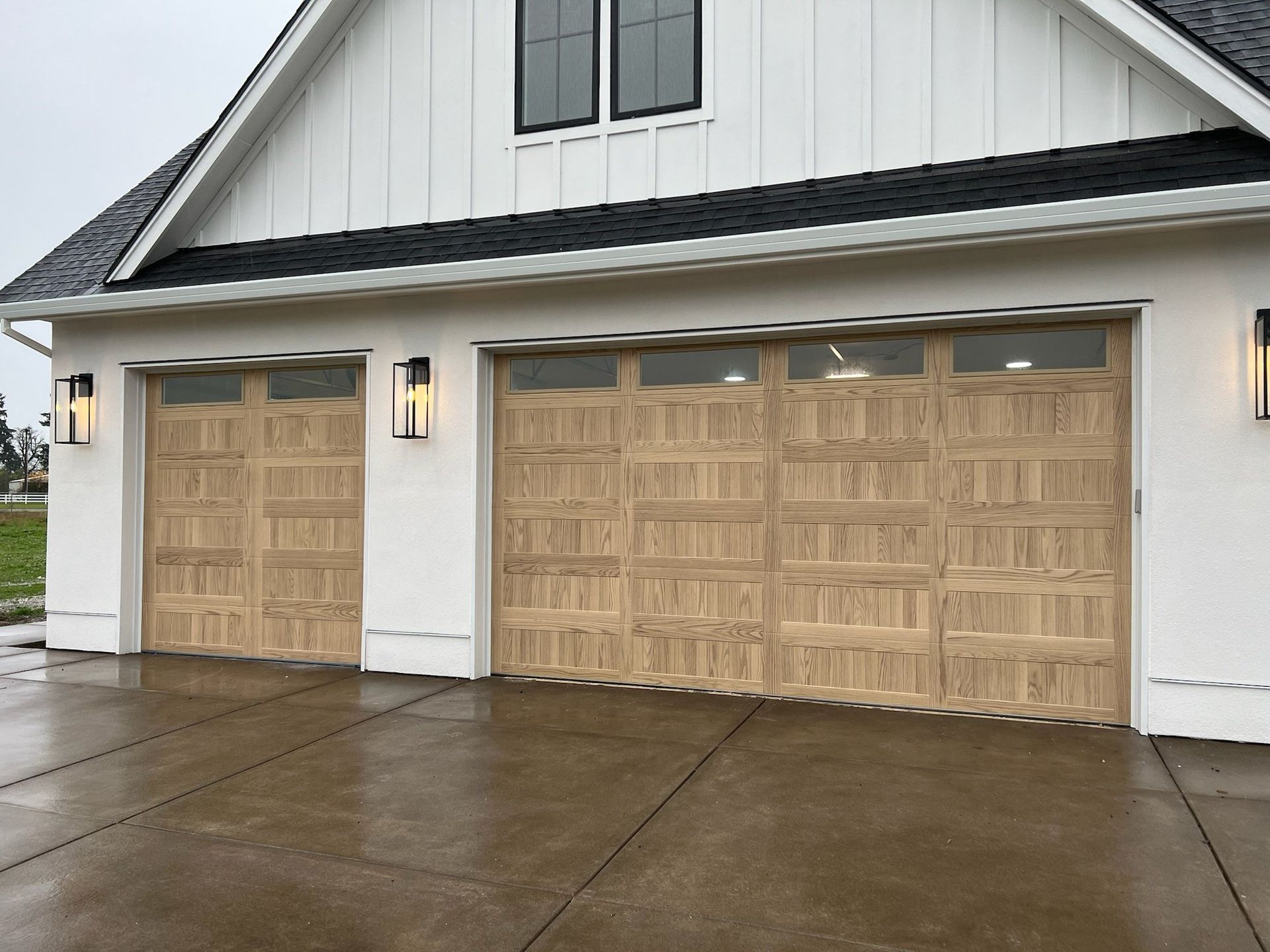 Tan garage doors with glass panels on a white house with black trim. Outdoor lighting and wet driveway.