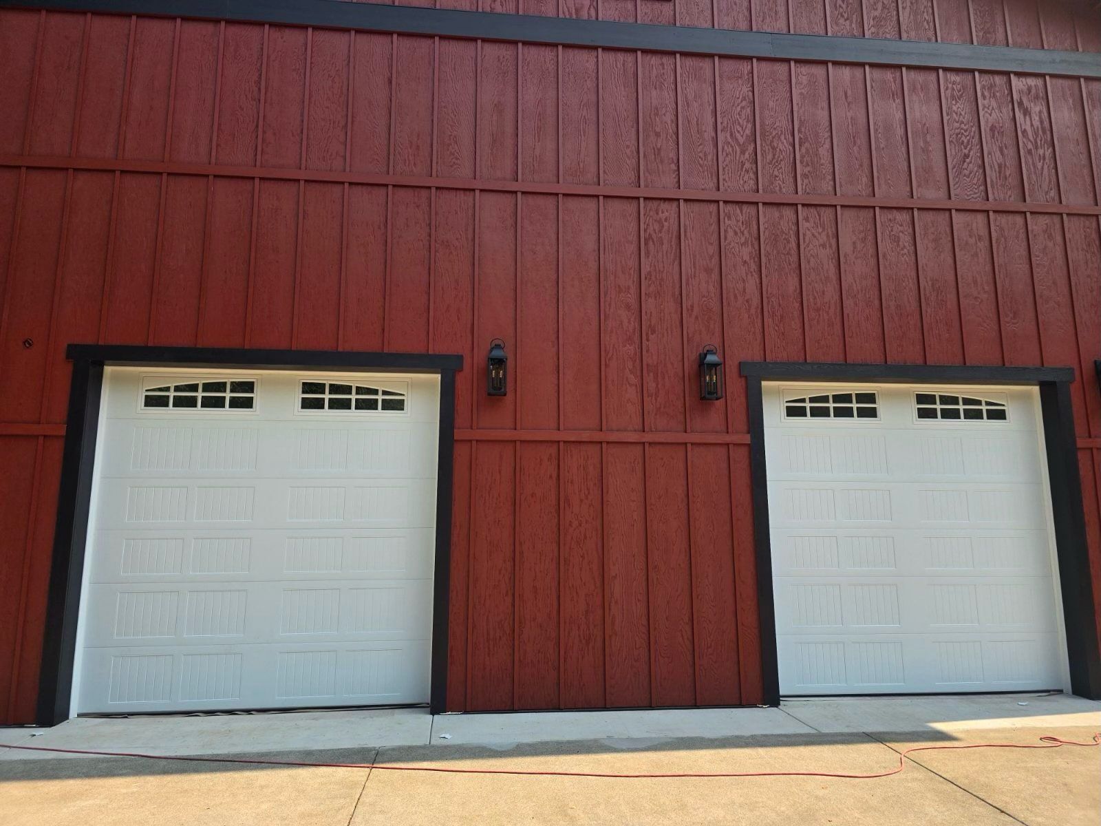 Two white garage doors on a red building with black trim, exterior lights, and a concrete driveway.
