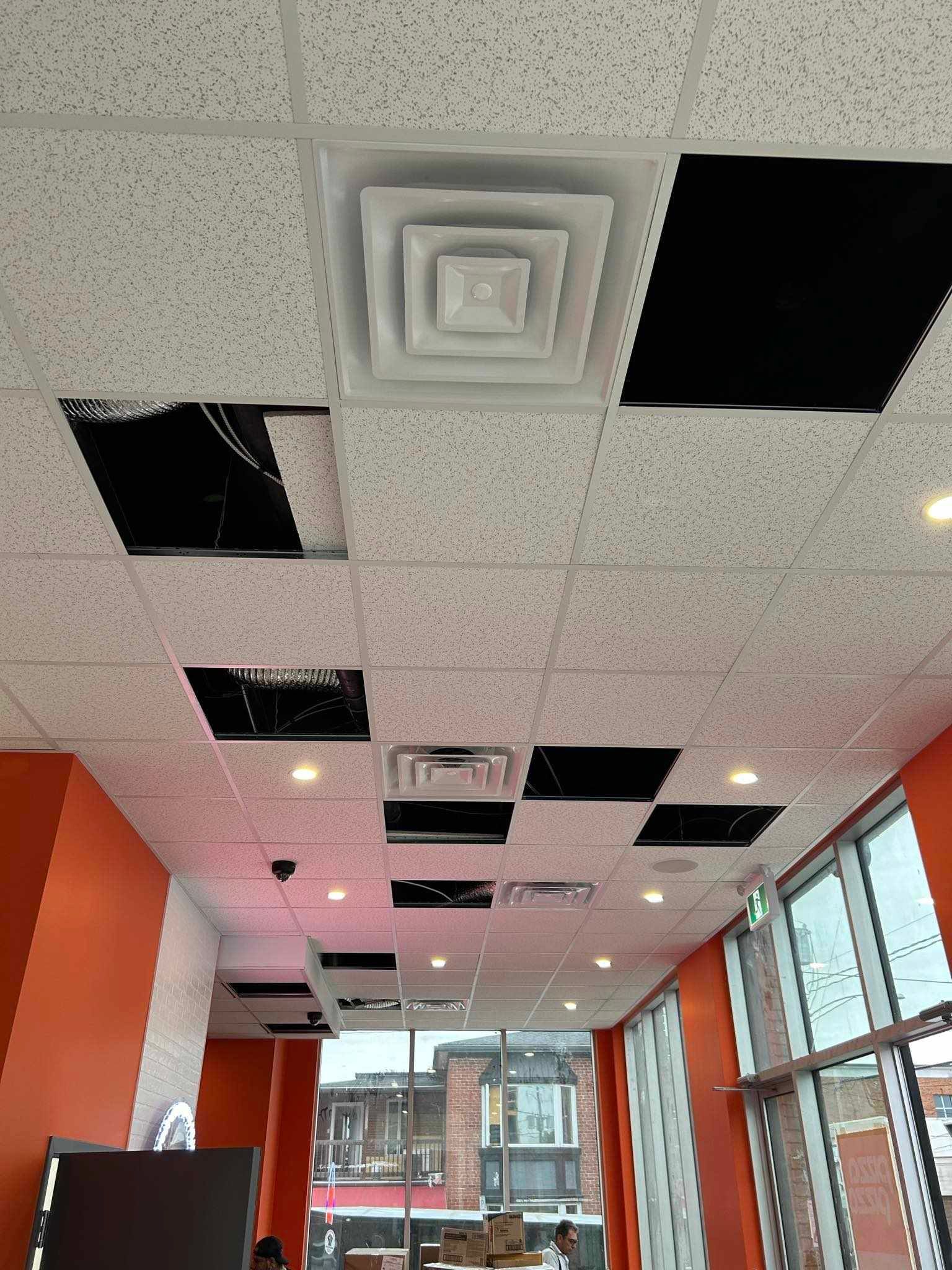 Plafond de bureau ou de hall d'entrée avec dalles carrées, panneaux noirs, grilles d'aération, murs orange et grandes fenêtres en dessous.