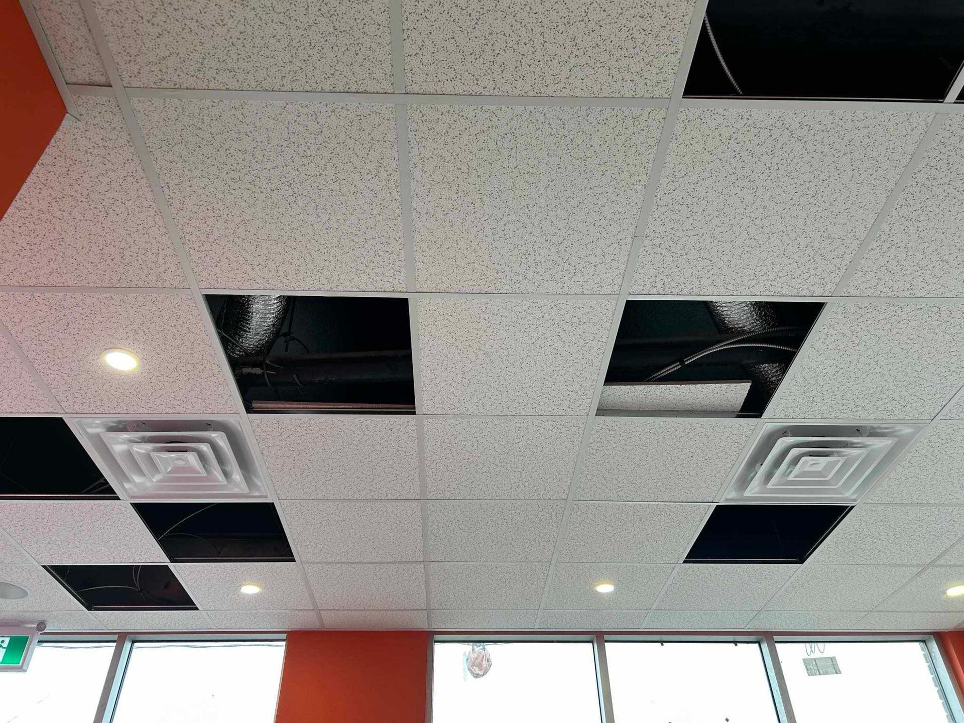 Plafond avec plusieurs dalles manquantes, aérations apparentes et une ligne de fenêtre en dessous.