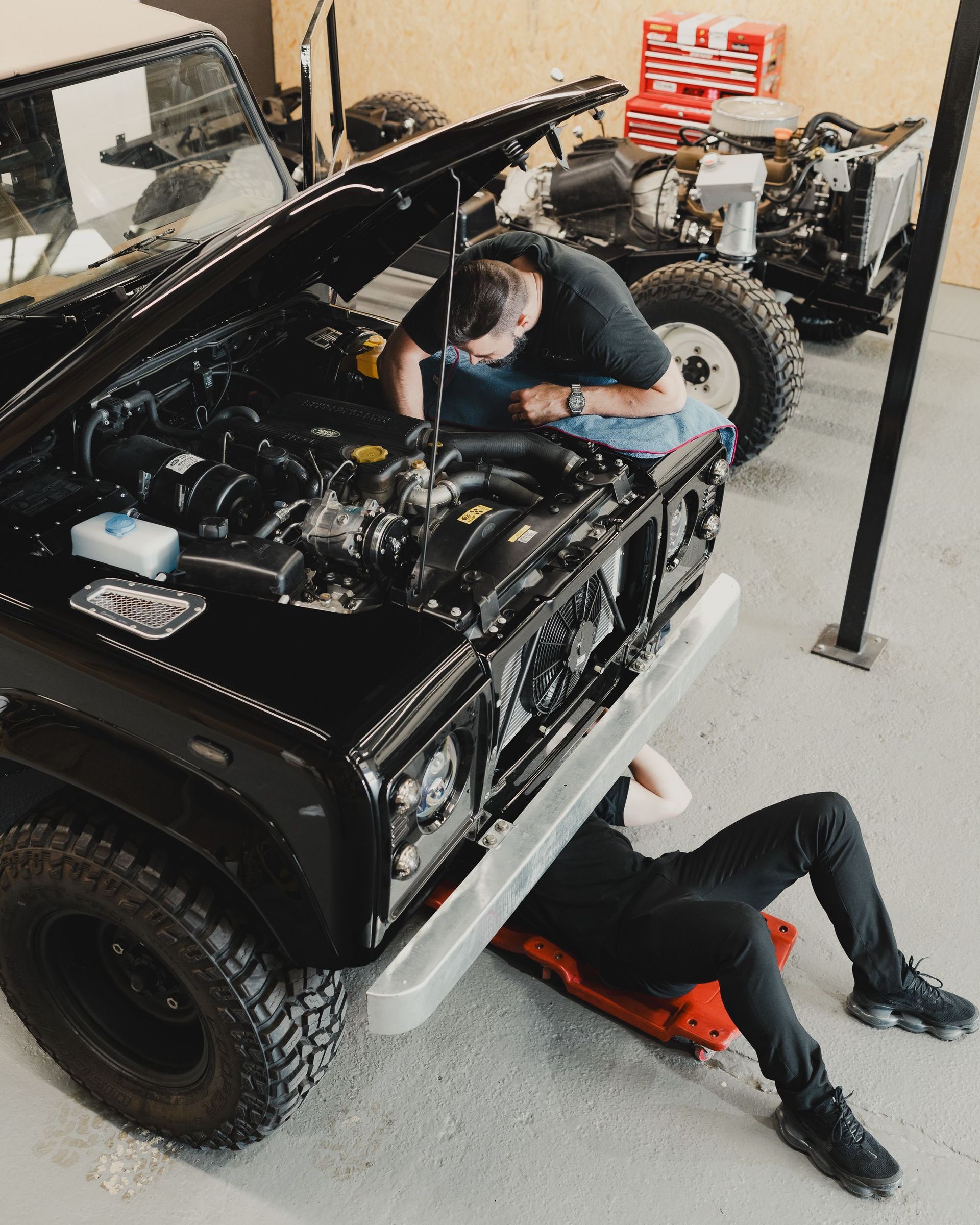 Two mechanics under a black off-road vehicle in a garage, working on the engine and underside.
